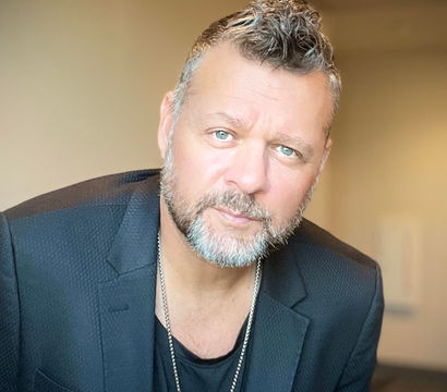 Confident indoor headshot of a middle-aged man with salt-and-pepper beard and short curly hair, wearing a black blazer and pendant necklace against a soft neutral background.