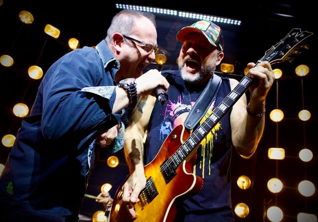 Two energetic rock musicians at a live concert—singer leaning into a shared mic and guitarist strumming a sunburst electric guitar amid warm vintage stage bulbs.