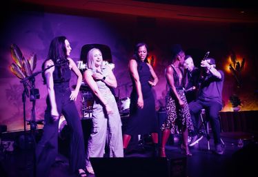 Five performers on a small nightclub stage — dancing singers and a guitarist playing live under purple-pink lighting.