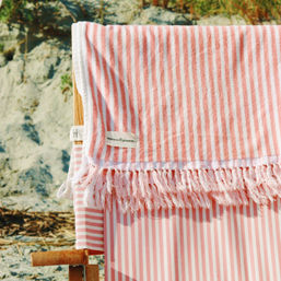 Pink-and-white striped beach towel with tassel fringe draped over a wooden beach chair on a sandy shore, summer coastal vibe.