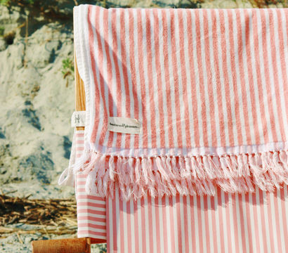 Pink-and-white striped beach towel with tassel fringe draped over a wooden beach chair on a sandy shore, summer coastal vibe.