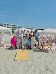 Group of women in colorful sun visors and summer outfits posing on a sandy oceanfront beach with pink-striped umbrellas, cabanas, folding chairs and a cornhole board under a clear blue sky near a coastal resort