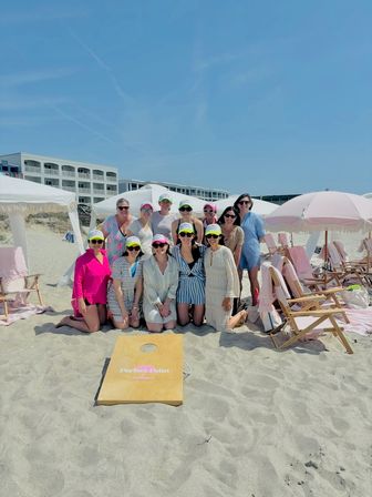 Group of women in colorful sun visors and summer outfits posing on a sandy oceanfront beach with pink-striped umbrellas, cabanas, folding chairs and a cornhole board under a clear blue sky near a coastal resort
