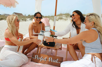 Four friends laughing and toasting with white tumblers under a striped beach umbrella on a sandy coastal dune — summer beach picnic with blankets, a small wooden table and portable speaker.