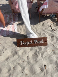 Wooden sign reading 'Perfect Point' on a sandy beach in front of pink-and-white striped beach chairs and towels