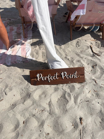 Wooden sign reading 'Perfect Point' on a sandy beach in front of pink-and-white striped beach chairs and towels