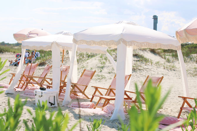 Sunny sandy beach with white fringed canopies and pink-striped wooden lounge chairs lined along coastal dunes, a white cooler in the foreground and a distant lighthouse under a blue sky.