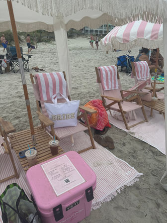Pink-and-white striped beach chairs and umbrella under a fringed canopy on a sandy beach, BRIDE tote on a chair, pink cooler, iced drinks and flip-flops — bachelorette beach setup