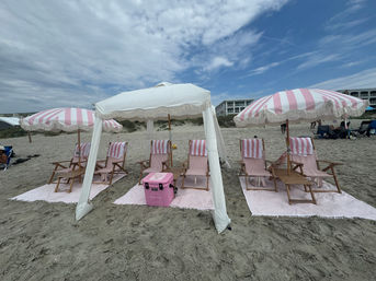 Playful pink-and-white striped umbrellas and wooden lounge chairs arranged under a white canopy on a sandy beach, with a pink cooler on matching striped mats, dunes and beachfront buildings under a partly cloudy sky.