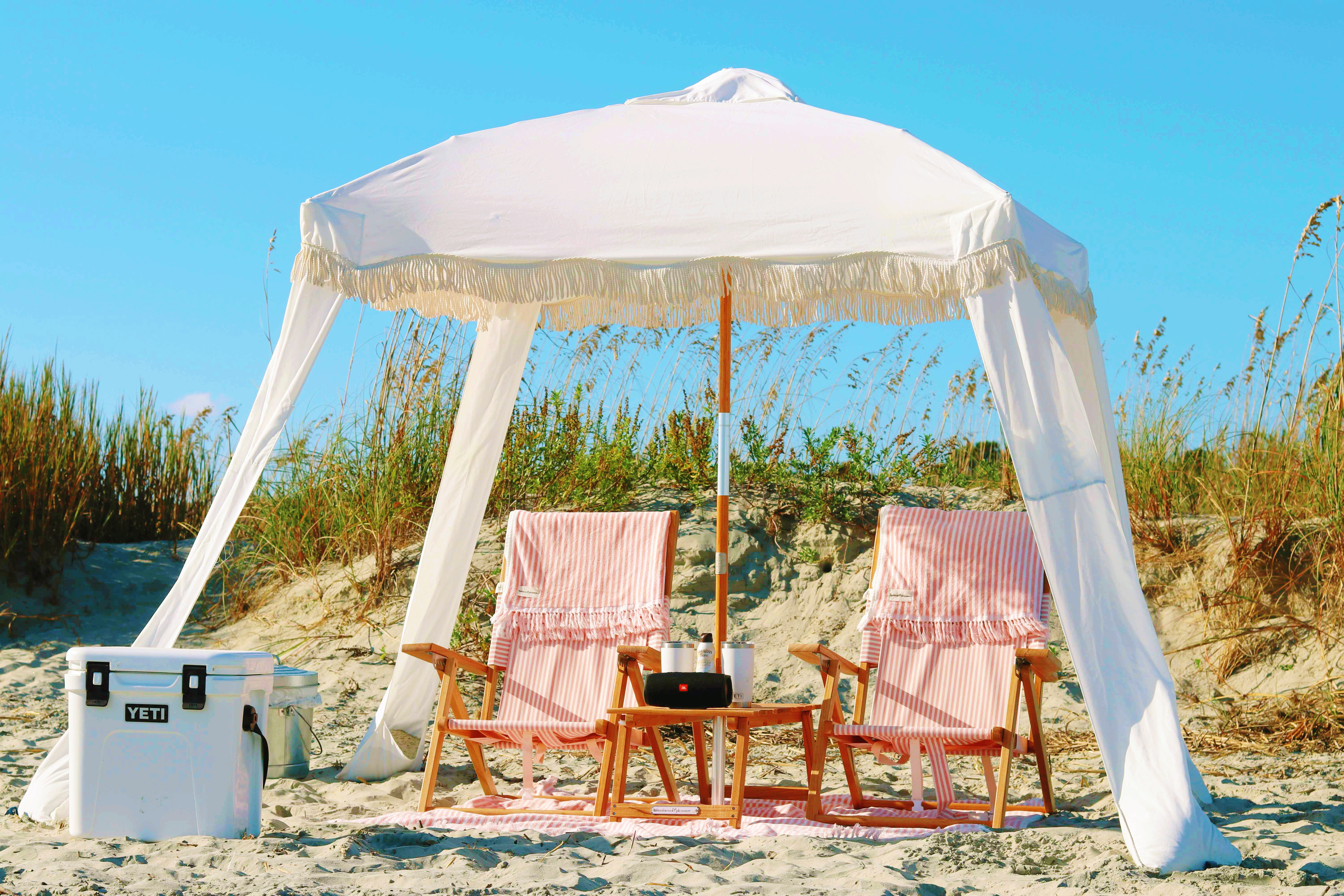 White fringe beach canopy shading two pink-striped wooden lounge chairs with a small table and white cooler on sandy coastal dunes under a bright blue sky.
