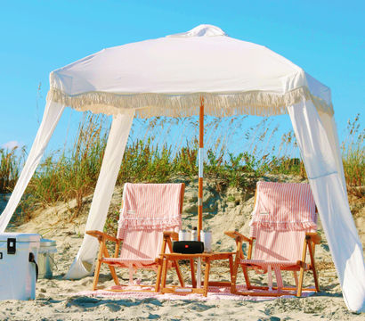 White fringe beach canopy shading two pink-striped wooden lounge chairs with a small table and white cooler on sandy coastal dunes under a bright blue sky.