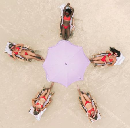 Aerial view of five sunbathers in red bikinis reclining on wooden lounge chairs arranged around a pink-and-white striped beach umbrella on pale sand.