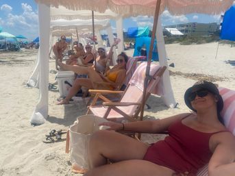 Group of women relaxing under fringe beach cabanas on a sunny sandy shoreline, reclining on striped lounge chairs with towels, coolers and colorful umbrellas for a summer beach day.