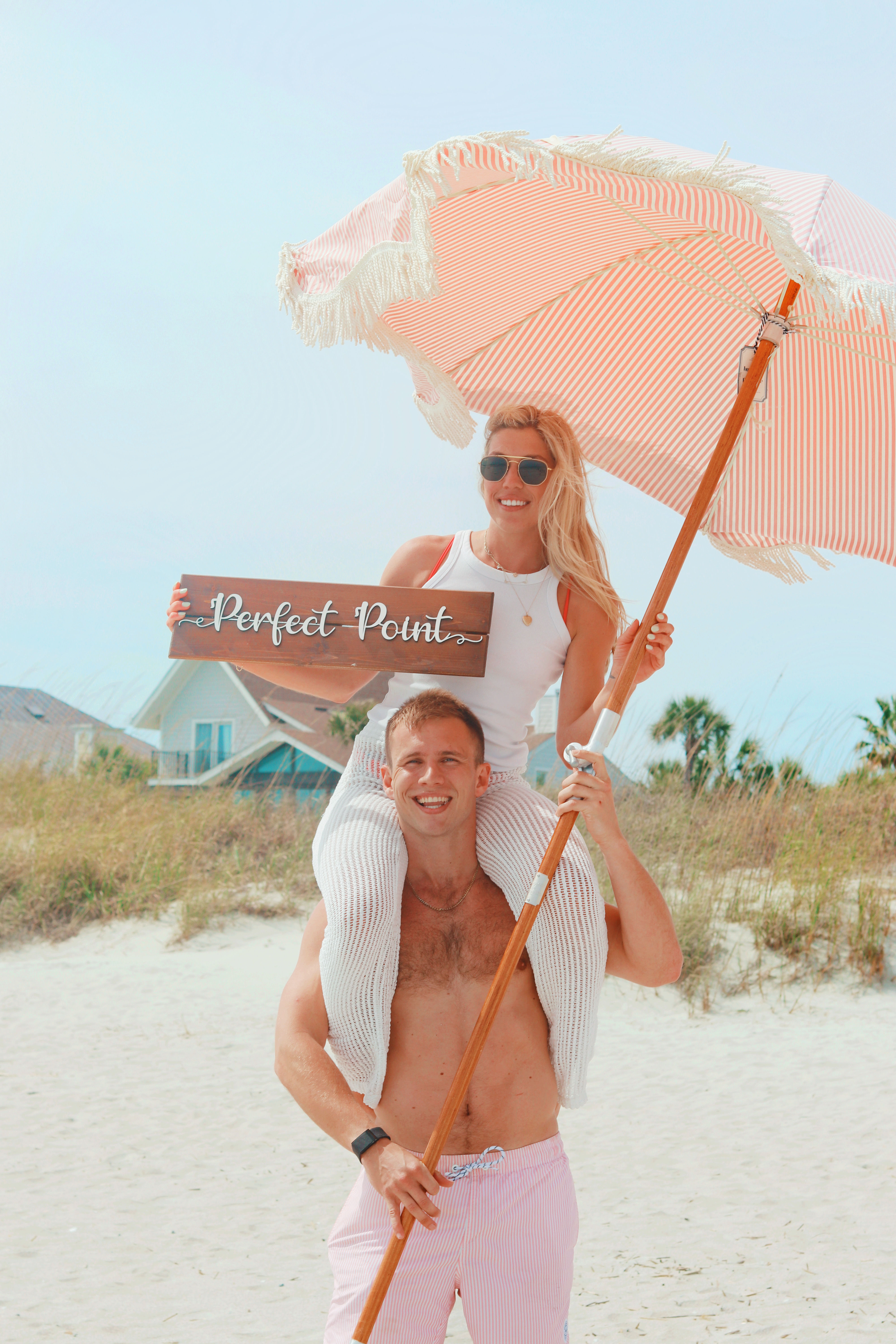 Smiling couple on a sunny sandy beach — woman on a man’s shoulders holding a pink striped beach umbrella and a wooden sign reading “Perfect Point,” with coastal dunes and beach houses in the background.