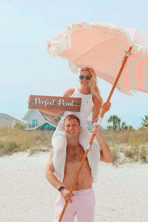 Smiling couple on a sunny sandy beach — woman on a man’s shoulders holding a pink striped beach umbrella and a wooden sign reading “Perfect Point,” with coastal dunes and beach houses in the background.