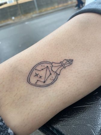 Whimsical minimal black-line tattoo of a small potion bottle with a crystal stopper containing a paper boat, dots and tiny stars on a person’s thigh