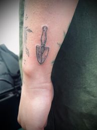 Close-up of an inner forearm minimal black linework tattoo of a small vintage garden trowel above the wrist, with faint botanical tattoos nearby.