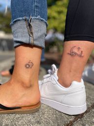 Two minimal ankle tattoos outdoors: left ankle shows a small sun with a crescent moon and stars above a cuffed blue jean and sandal; right ankle features a simple wave with a crescent and star above black leggings and a white sneaker on concrete.