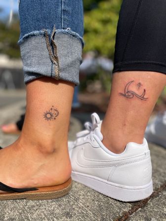 Two minimal ankle tattoos outdoors: left ankle shows a small sun with a crescent moon and stars above a cuffed blue jean and sandal; right ankle features a simple wave with a crescent and star above black leggings and a white sneaker on concrete.