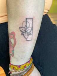 Minimal black linework flower tattoo overlaying a vertical rectangle and dotted semicircle on an inner forearm; colorful heart tattoo and woven bracelets visible near the wrist.