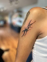Close-up of an upper-arm black tribal-style tattoo near the shoulder with a small floral outline above, person wearing a white tank top, soft-focus indoor background