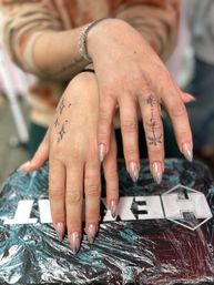 Close-up of hands on a plastic-covered surface showcasing chrome stiletto manicure with tiny rhinestones and delicate black line hand tattoos — star cluster on one hand and a sun-and-palm motif on a finger, nail art and fresh manicure style.