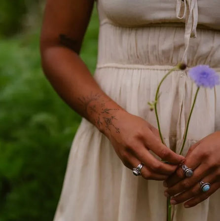 Person in a cream sundress holding two purple wildflowers in a green meadow, showing a delicate forearm tattoo and silver turquoise rings.
