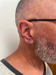 Close-up of a right earlobe with a tiny triangle-in-circle tattoo, black glasses arm, short buzzed hair and salt-and-pepper beard against a white wall.