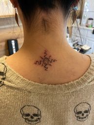 Close-up of nape showing a small black line tattoo — central spiral sun with four wavy rays on the back of the neck, person wearing a beige skull-patterned sweater.
