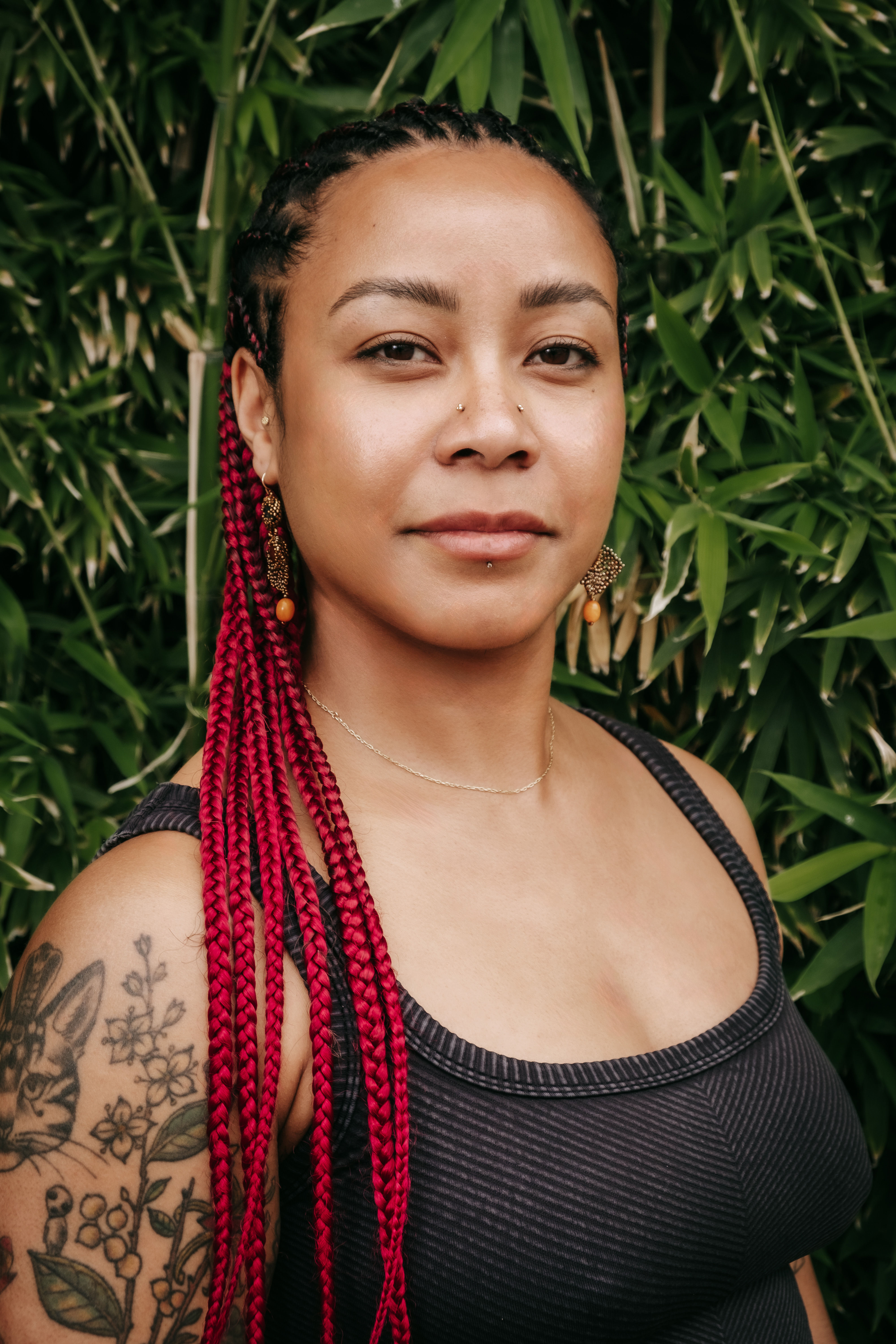 Close-up outdoor portrait of a person with bold red braids, nose and lip piercings, gold-and-orange dangling earrings, and a floral rabbit tattoo on the upper arm, wearing a black tank top against a green bamboo backdrop.