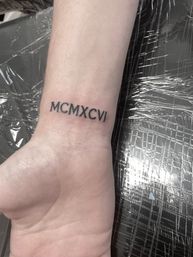 Close-up of a black-ink Roman numerals wrist tattoo reading MCMXCVI on the inner wrist, fresh-looking on pale skin against a plastic-covered surface.