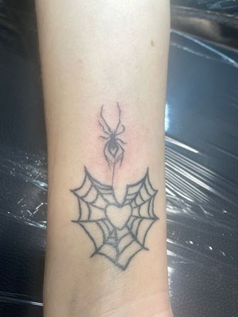 Close-up of an inner forearm/wrist black-ink tattoo: a small spider dangling above a heart-shaped spiderweb, simple linework
