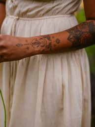 Close-up of a person's forearm in a cream linen dress outdoors, showcasing black linework tattoos: crescent moon, delicate leaf motifs, dotted accents and a floral mandala near the elbow.