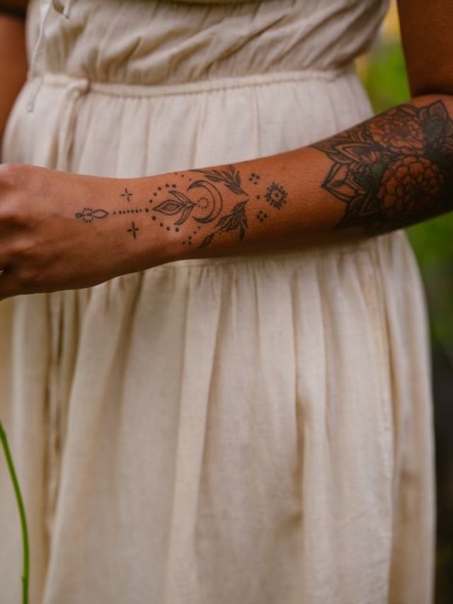Close-up of a person's forearm in a cream linen dress outdoors, showcasing black linework tattoos: crescent moon, delicate leaf motifs, dotted accents and a floral mandala near the elbow.