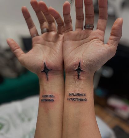 Close-up of matching wrist tattoos on two forearms: black four-point star near each palm and text 'CONTROL NOTHING' (left) and 'INFLUENCE EVERYTHING' (right), fresh ink