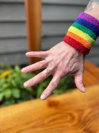 Close-up of a hand wearing a rainbow-striped wristband, displaying a small zigzag finger tattoo against a blurred wooden deck and green planter background.