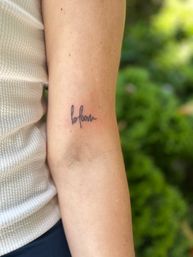 Small cursive "bloom" tattoo on inner forearm near elbow, person in white ribbed top photographed outdoors with blurred green garden background