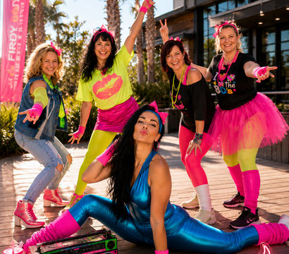 Five women in neon 1980s-inspired workout outfits posing on a sunny palm-lined boardwalk — one in metallic blue doing a split by a pink boombox, others in bright tutus, leg warmers and graphic tees, smiling and striking energetic dance poses.
