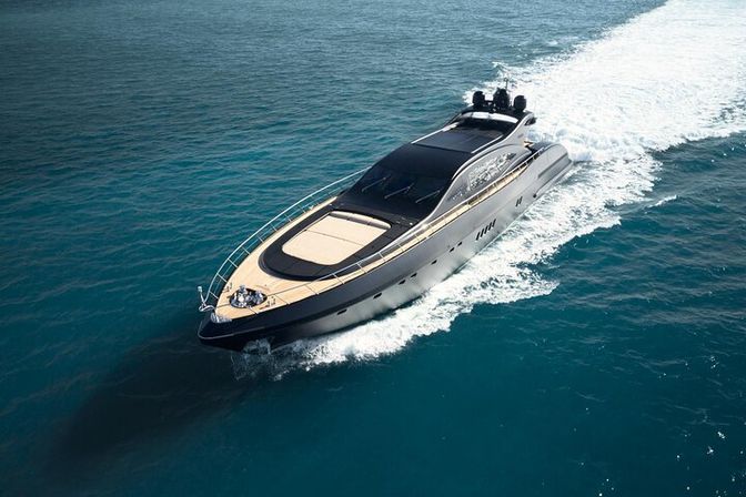 Aerial view of a sleek black luxury motor yacht cutting through deep blue sea, sunlit foredeck with cushioned seating and a white foamy wake trailing behind.