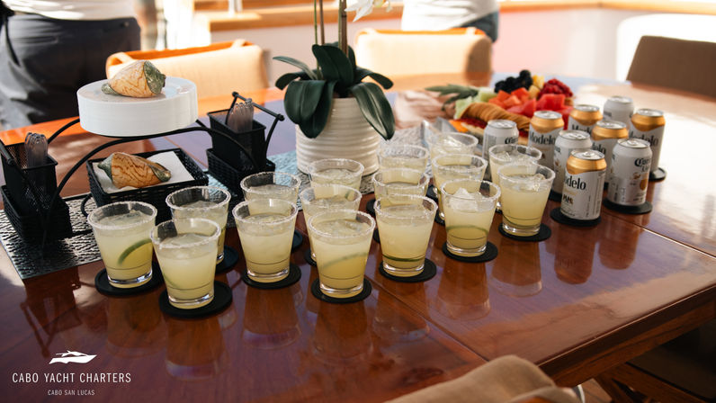 Sunlit yacht deck table in Cabo San Lucas with rows of salted-rim margaritas, canned beers, a fruit platter and orchid centerpiece.