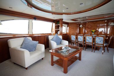 Sunlit luxury yacht salon with cream armchairs and blue cushions, glass-top coffee table, polished wood paneling and a dining area with patterned chairs beside large ocean-facing windows.