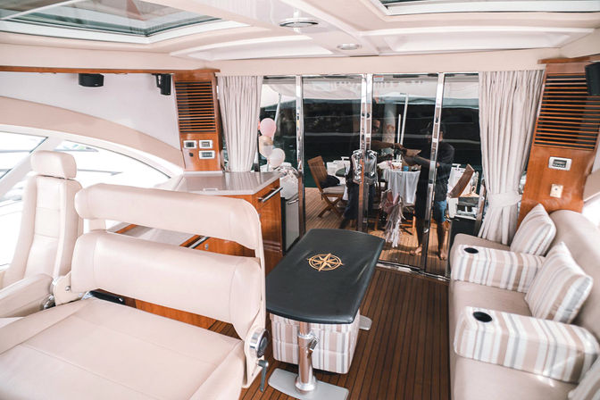 Onboard luxury yacht interior with cream leather seats, striped sofa with cup holders, teak decking, black bench with compass motif and glass sliding doors to an aft dining area with wooden chairs and balloons
