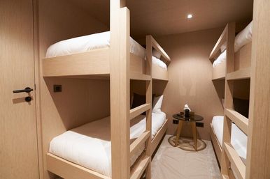 Cozy compact room with two sets of light-wood bunk beds (four beds), crisp white bedding, a small round bedside table with tissues and bottle, and warm recessed lighting.