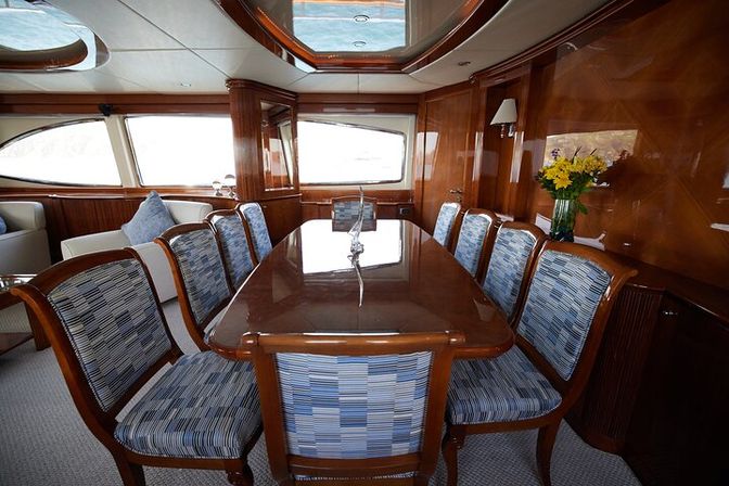 Polished teak dining table surrounded by blue-striped upholstered chairs in a sunlit luxury yacht salon with glossy wood paneling, panoramic windows and a vase of yellow flowers.