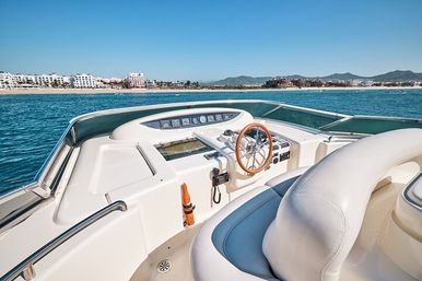 Sun-drenched luxury yacht helm with wooden steering wheel and cushioned seat, turquoise ocean and sandy beachfront resorts with distant mountains under a clear blue sky
