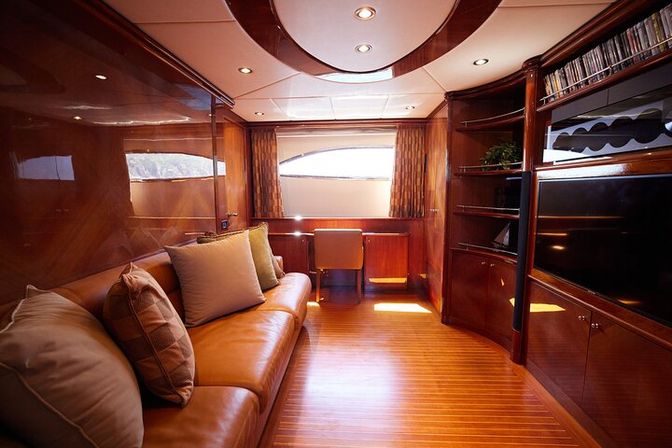 Luxury yacht salon interior with glossy mahogany paneling and teak floor, tan leather sofa with pillows, built-in shelving and flat-screen TV, and a small desk beneath an oval window flooding the space with natural light.