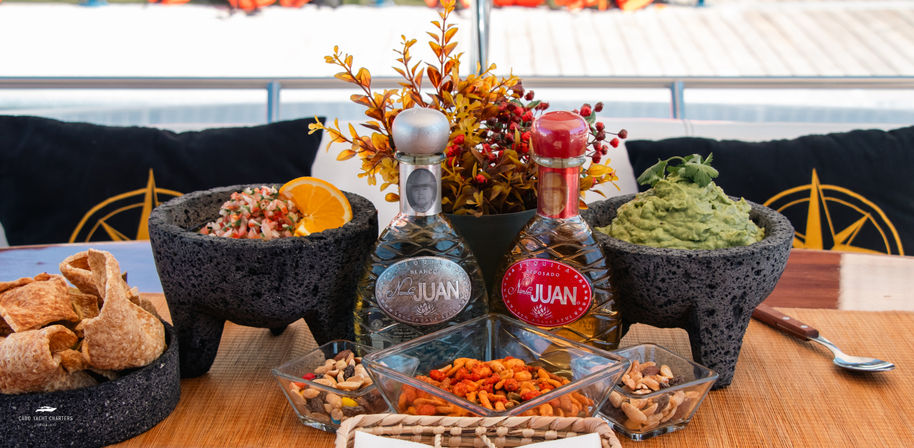 Mexican appetizer spread on a yacht table: guacamole and pico de gallo in volcanic stone bowls, tortilla chips, seasoned nuts and two decorative tequila bottles.