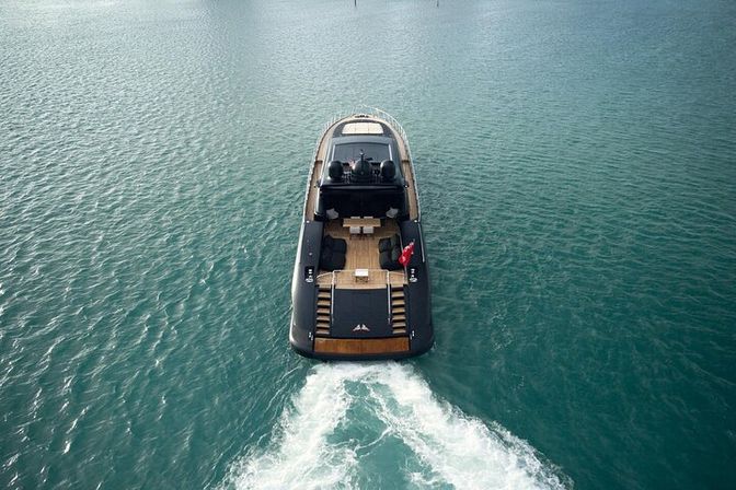 Aerial view of a sleek black luxury motor yacht cruising through calm turquoise water, stern cutting a white foamy wake.