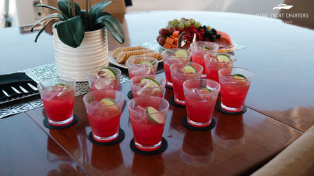 Row of pink lime‑garnished cocktails on coasters arranged on a glossy wooden table aboard a yacht, with a potted plant and a fruit and cracker platter in the background