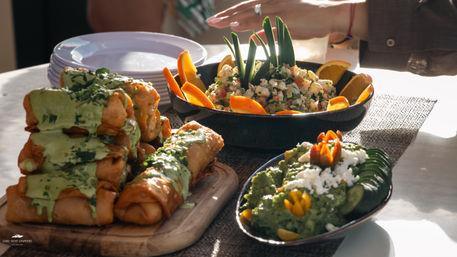 Sunlit table of Mexican appetizers: stacked crispy flautas drizzled with green salsa, a bowl of citrus shrimp ceviche garnished with orange wedges, and a plate of guacamole topped with queso fresco and cucumber slices.